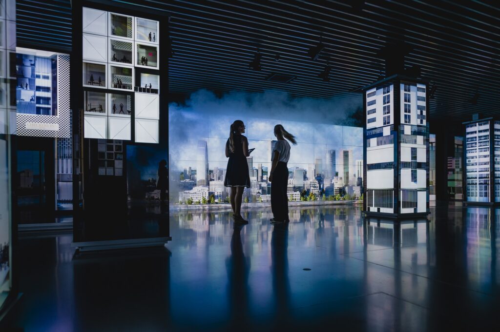 Two young women standing in a dark room with big screens which show futuristic buildings