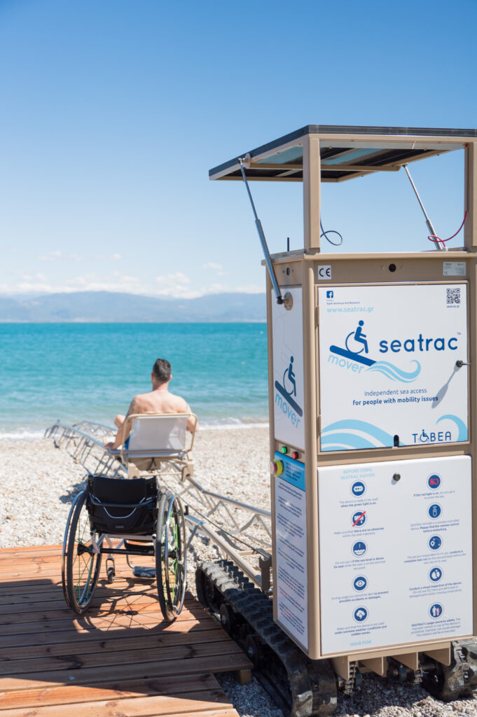 SEATRAC Strandzugangssystem am Strand aufgebaut; im Hintergrund sieht man eine Person, die vorher im Rollstuhl saß, wie sie sich auf einem Sitz auf Schienen Richtung Meer bewegt