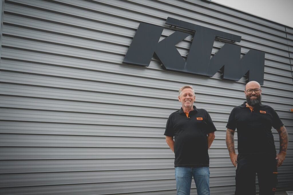 Zwei Projektpartner von KTM stehen vor der Werkshalle mit KTM-Logo im Hintergrund