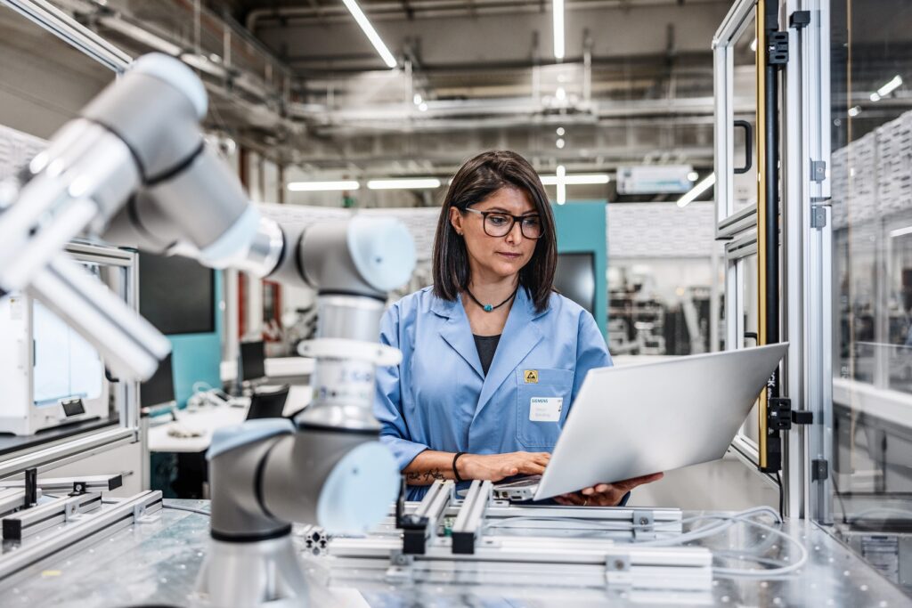 Eine Frau steht in einer industriellen Produktion vor einem Roboterarm mit einem aufgeklappten Laptop in der Hand, auf den sie schaut