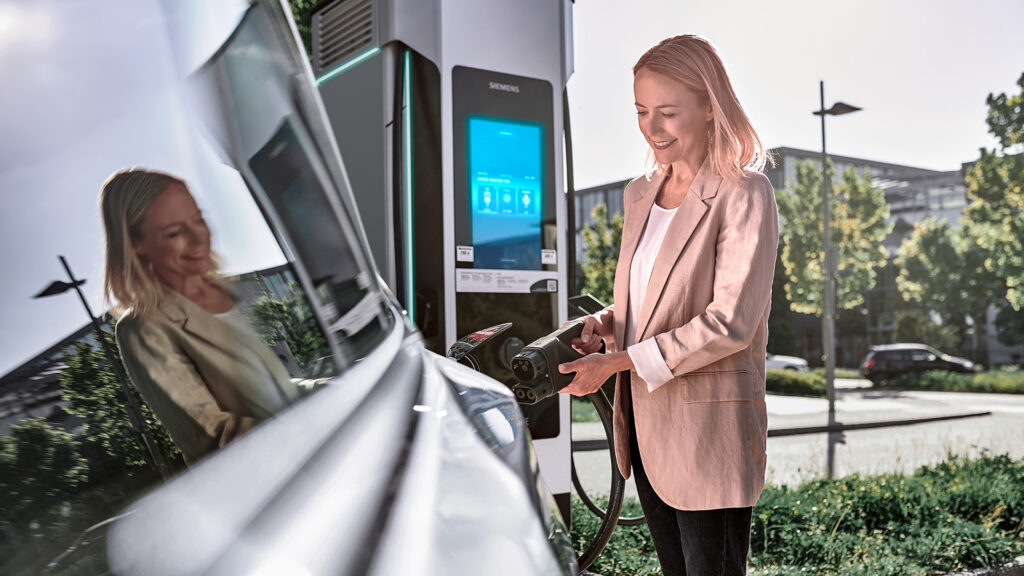 Eine Frau ist dabei, den Stecker in ein Elektroauto zu stecken, der von einer Siemens-Ladesäule abgeht.