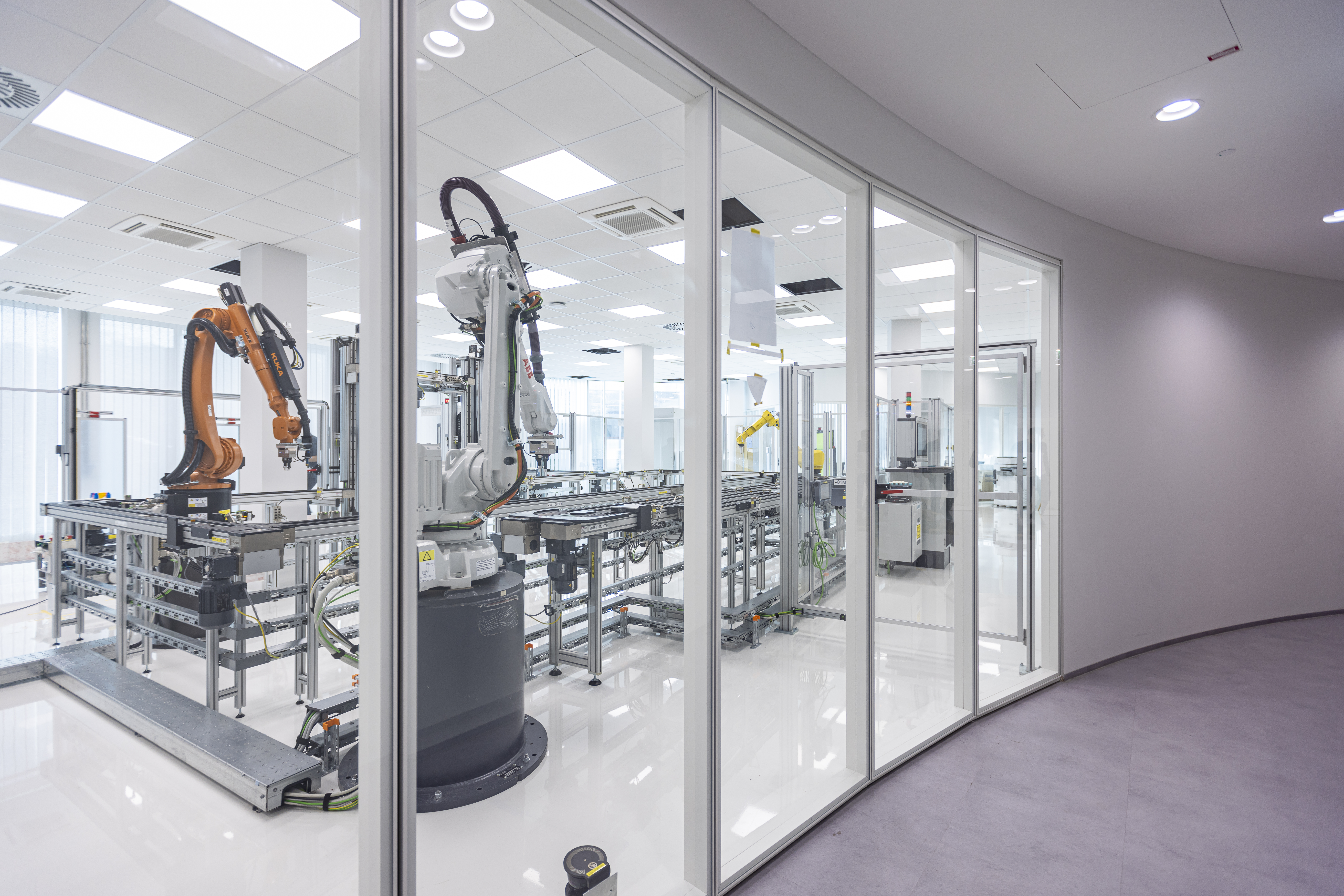 Das Bild zeigt das Manufactoring Solutions Lab am neu eröffneten Standort Linz. Hinter einer Glasfront sind zwei Greifarme - einer orange, einer weiß schwarz zu sehen.